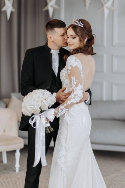 Bir gelin ve damat sandalyenin önünde fotoğraf için poz veriyorlar. Gelin beyaz bir elbise giyiyor ve damat siyah bir takım elbise giyiyor. Gelin elinde bir buket çiçek tutuyor.