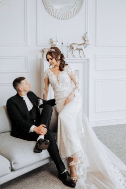 Bir gelin ve damat bir kanepede oturmuş resim için poz veriyorlar. Gelin uzun beyaz bir elbise giyiyor ve damat bir takım elbise giyiyor. Sahne mutlu ve romantik.