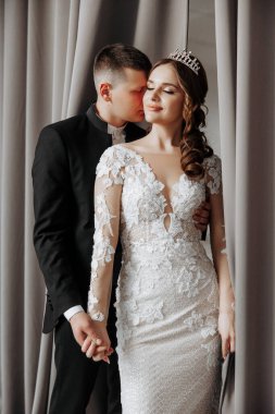 Bir gelin ve damat bir düğün töreninde birbirlerini kucaklıyorlar. Gelin, başında taç olan beyaz bir elbise giyiyor. Adam takım elbise ve kravat takıyor. Sahne samimi ve romantik.