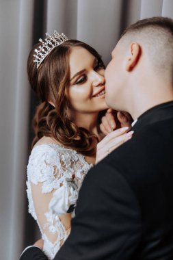 Bir gelin ve damat birbirlerinin dudaklarını öpüyor. Gelin taç takıyor ve damat da takım elbise giyiyor. Sahne romantik ve mutlu.