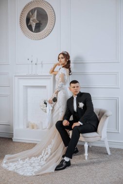 Gelin ve damat şöminenin önünde fotoğraf için poz veriyorlar. Gelin beyaz bir elbise giyiyor ve damat siyah bir takım elbise giyiyor. Sahne romantik ve samimi.