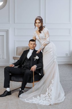 Gelin ve damat, arka planda sandalye olan bir odada fotoğraf için poz veriyor. Gelin uzun beyaz bir elbise giyiyor ve damat bir takım elbise giyiyor. Sahne mutlu ve kutlama dolu.