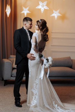 Gelin ve damat bir kanepenin önünde, gelin bir buket tutuyor. Çiftler düğünlerini kutlarken sahne romantik ve mutlu.