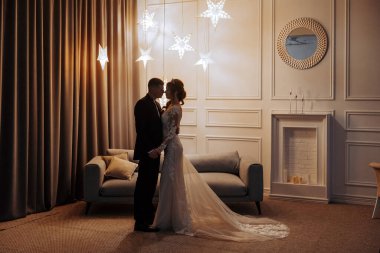 Bir gelin ve damat bir kanepede ve şömineli bir odada duruyorlar. Sahne romantik ve samimi.