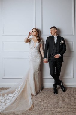 Bir gelin ve damat beyaz bir duvarın önünde poz veriyorlar. Gelin uzun beyaz bir elbise giyiyor ve damat siyah bir takım elbise giyiyor. Sahne mutlu ve kutlama dolu.