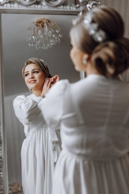 Bir kadın düğün için hazırlanıyor ve aynada kendine bakıyor. Beyaz bir elbise giyiyor ve saçları topuz şeklinde.