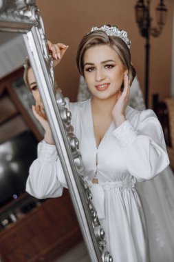 Beyaz elbiseli bir kadın aynanın önünde duruyor. Gülümsüyor ve yansımasına bakıyor.