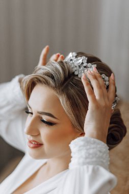 Bir kadın düğün için hazırlanıyor ve taç takıyor. Beyaz bir elbise giyiyor ve saçları topuz şeklinde.