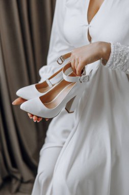 Bir kadın bir çift beyaz ayakkabı tutuyor. Ayakkabılar yüksek topuklu ve büyük ihtimalle düğün gibi özel durumlar için. Kadın, ayakkabıları tamamlayan beyaz bir elbise giyiyor.