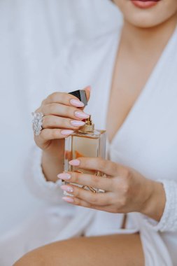 Bir kadın bir şişe parfüm tutuyor ve beyaz bir elbise giyiyor. Parfüm şişesi altın renginde ve pembe bir bluzu var. Kadının elleri ojeyle süslenmiş ve yüzünde bir gülümseme var.