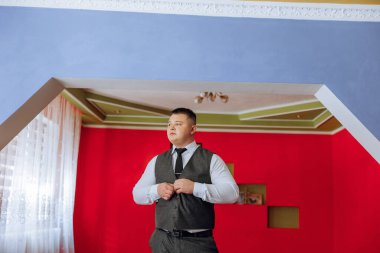 Takım elbiseli ve kravatlı bir adam kırmızı bir duvarın önünde duruyor. Kravatını düzeltiyor ve kameraya bakıyor.