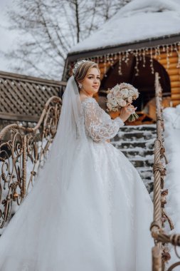 Beyaz gelinlikli bir kadın bir binanın önündeki merdivende dikiliyor. Elinde bir buket çiçek tutuyor.