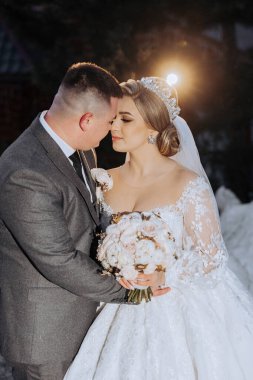 Bir gelin ve damat ellerinde bir buket çiçekle birbirlerini kucaklıyorlar. Gelin uzun beyaz bir elbise giyiyor ve damat bir takım elbise giyiyor. Sahne romantik ve mutlu.