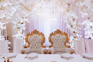 Masanın her iki tarafında birer sandalye bulunan beyaz ve altın kaplamalı bir oda. Sandalyeler çiçeklerle süslenir ve çiçek desenli bir duvarın önüne yerleştirilir.