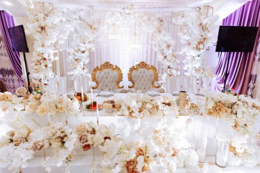 Ortasında büyük bir masa olan beyaz ve altın dekore edilmiş bir oda. Masa beyaz çiçeklerle kaplı ve iki sandalyesi var. Ayrıca duvarda iki televizyon var, biri solda, biri sağda.