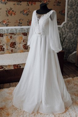 Çiçek desenli uzun beyaz bir elbise bir odada sergilenir. Elbise parlak malzemeden yapılmıştır ve uzun bir treni vardır.