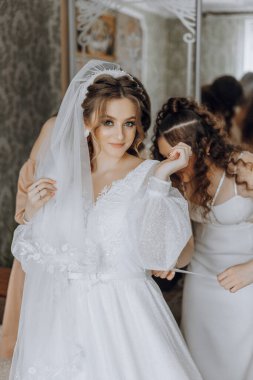 Bir gelin, duvağını düzeltmeye yardım eden bir kadınla düğününe hazırlanıyor. Gelin beyaz bir elbise ve duvak takıyor ve kadın elinde bir kurdele tutuyor.