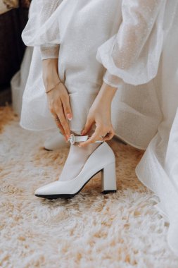 Beyaz elbiseli bir kadın düğün ayakkabılarını giymeye hazırlanıyor. Kayışları ayarlıyor ve güvenli olduklarından emin oluyor. Yaklaşan düğün için beklenti ve heyecan kavramı