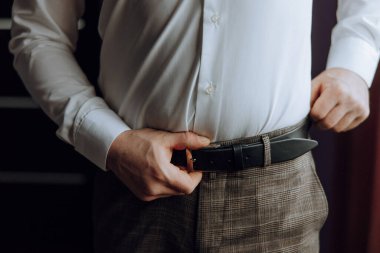 Bir adam beyaz gömlek giyerken kemerini ayarlıyor. Formalite anlayışı ve detaylara dikkat, adam kemerinin düzgün bağlandığından emin olmak için zaman ayırıyor.