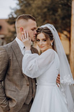 Bir gelin ve damat fotoğraf için poz veriyor. Gelin beyaz bir elbise giyiyor ve damat bir takım elbise giyiyor. İkisi de gülümsüyor ve birbirlerine sarılıyorlar. Sahne mutlu ve sevgi dolu.