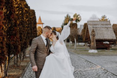 Bir gelin ve damat binanın önünde öpüşüyor. Gelin bir buket tutuyor.