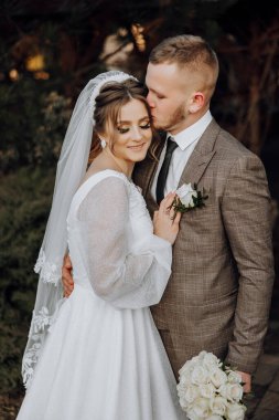 Gelin ve damat birbirine sarılıyor, gelin beyaz bir elbise ve damat takım elbise giyiyor.