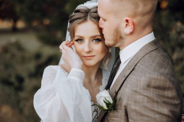 Bir gelin ve damat ormanda birbirlerine sarılıyorlar. Gelin beyaz bir elbise giyiyor ve damat da takım elbise giyiyor.