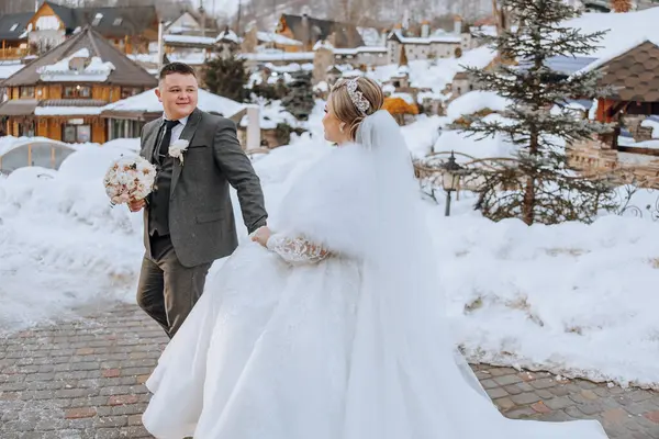 Gelin ve damat karda yürüyorlar, gelin kürk şort giyiyor. Gelin elinde bir buket çiçek tutuyor.