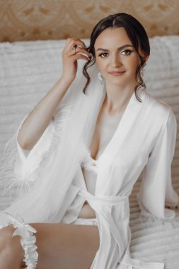 Beyaz cüppeli bir kadın bir yatakta oturuyor. Duvak takmış ve saçları topuz gibi olmuş.