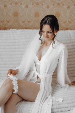 Beyaz elbiseli bir kadın bacak bacak üstüne atmış bir kanepede oturuyor. Beyaz dantelli sütyen ve beyaz bir peçe takıyor.