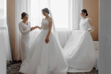 Üç kadın düğün için hazırlanıyor. Biri beyaz bir elbise giyiyor ve diğer ikisi de beyaz elbiseler giyiyor. Gelin, annesi ve kız kardeşiyle hazırlanıyor.