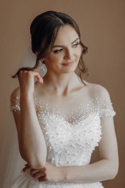 Gelinlik giymiş bir kadın gülümsüyor ve kameraya bakıyor. İnci bir kolye ve küpe takıyor.