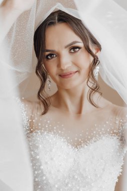 Beyaz bir elbise ve duvak takan bir kadın var. Gülümsüyor ve kameraya bakıyor.