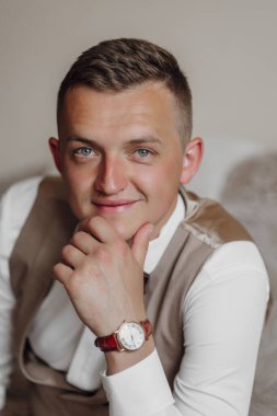 Yelek ve saat giyen bir adam kanepede oturuyor. Gülümsüyor ve kameraya bakıyor.