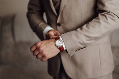Takım elbiseli bir adam saat takıyor ve ceketini ayarlıyor. Saat kırmızı ve beyaz.