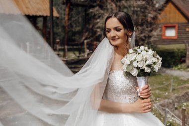 Bir gelin, beyaz bir duvak ve bir buket beyaz çiçekle bir binanın önünde duruyor.