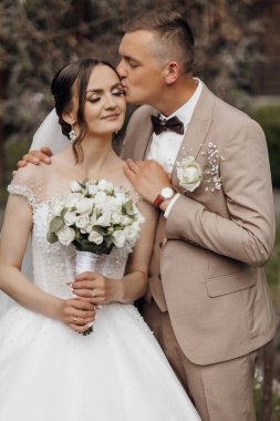 Bir gelin ve damat bir buket beyaz çiçek tutarken birbirlerini öpüyorlar. Gelin beyaz bir elbise giyiyor ve damat da takım elbise giyiyor.