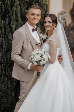 Bir gelin ve damat bir çalılığın önünde fotoğraf için poz veriyorlar. Gelin beyaz bir elbise giyiyor ve damat bir takım elbise giyiyor. Ellerinde çiçekler tutuyorlar.