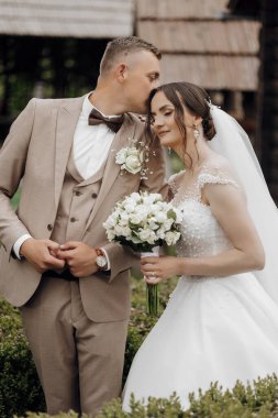 Gelin ve damat bir resim için poz veriyor, gelin elinde bir buket beyaz çiçek tutuyor. Adam takım elbise ve kravat takıyor ve kadın beyaz bir elbise giyiyor. Sahne mutlu ve romantik.
