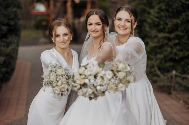 Beyaz elbiseli üç kadın çiçekli bir resim için poz veriyor. Gelin elinde bir buket beyaz çiçek tutarken diğer iki kadın daha küçük buketler tutuyor.