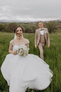 Bir gelin ve damat düğün günlerinde bir tarlada yürüyorlar. Gelin bir buket çiçek tutuyor ve damat takım elbise giyiyor. Sahne mutlu ve kutlama dolu.