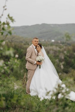 Bir gelin ve damat çimenli bir yamaçta durup resim için poz veriyorlar. Gelin beyaz bir elbise giyiyor ve damat bir takım elbise giyiyor. Bir buket çiçek ve çiçek tutuyorlar.