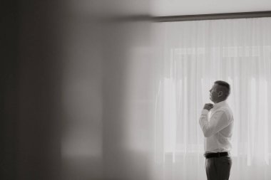 Bir adam kravatını bağlamaya hazırlanıyor. O bir pencerenin önünde duruyor ve oda loş bir şekilde aydınlanıyor. Adam görevine odaklanmış durumda ve görüntünün genel havası ciddi ve odaklanmış durumda.