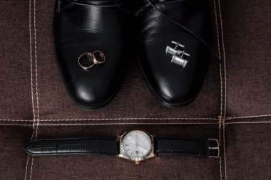 Bir adamın ayakkabıları ve saati kahverengi bir kanepede sergilenir. Saat siyah bantlı gümüş bir saat. Ayakkabılar siyah ve sol ayağında altın bir yüzük var. Zarafet ve incelik kavramı