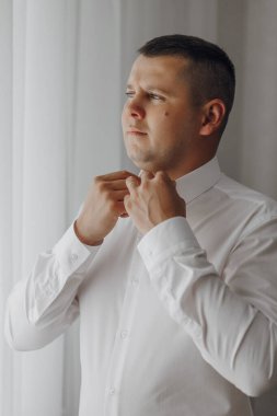 Bir adam beyaz gömlek giymeye hazırlanıyor. Yakasını ve kollarını düzeltiyor.