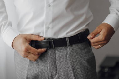 Beyaz gömlek ve gri pantolon giyen bir adam var. O, kemerini ayarlıyor. Formalite kavramı ve detaylara dikkat