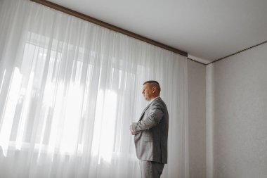 Takım elbiseli bir adam pencerenin önünde duruyor, kravatını düzeltiyor. Oda sade ve beyaz. Geniş bir penceresi var.