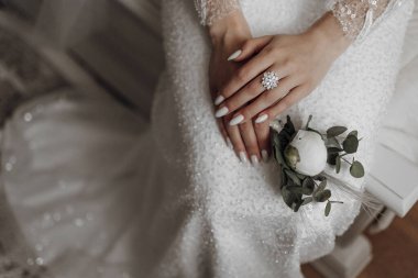 Bir kadın elleri kucağında bir sandalyede oturuyor. Beyaz bir elbise ve elmas bir yüzük giyiyor.