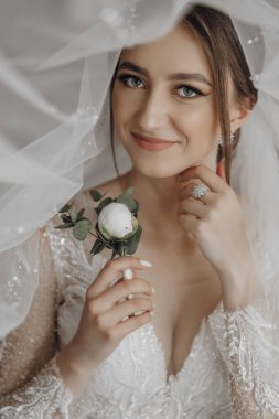 Bir kadın beyaz bir elbise giyiyor ve elinde bir çiçek tutuyor. Gülümsüyor ve kameraya bakıyor.