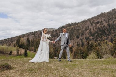 Gelin ve damat, arka planda dağların güzel manzarası olan bir tarlada birlikte yürüyorlar.